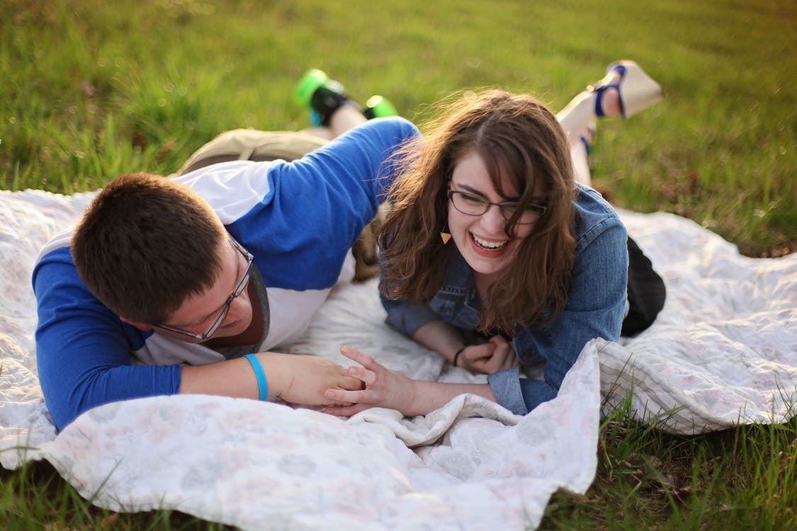 A couple laughing while holding hands on the blanket on top of the grass.