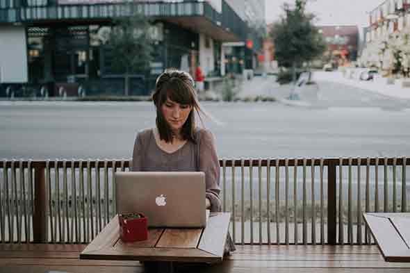 A woman sitting on a wooden bench facing a laptop
